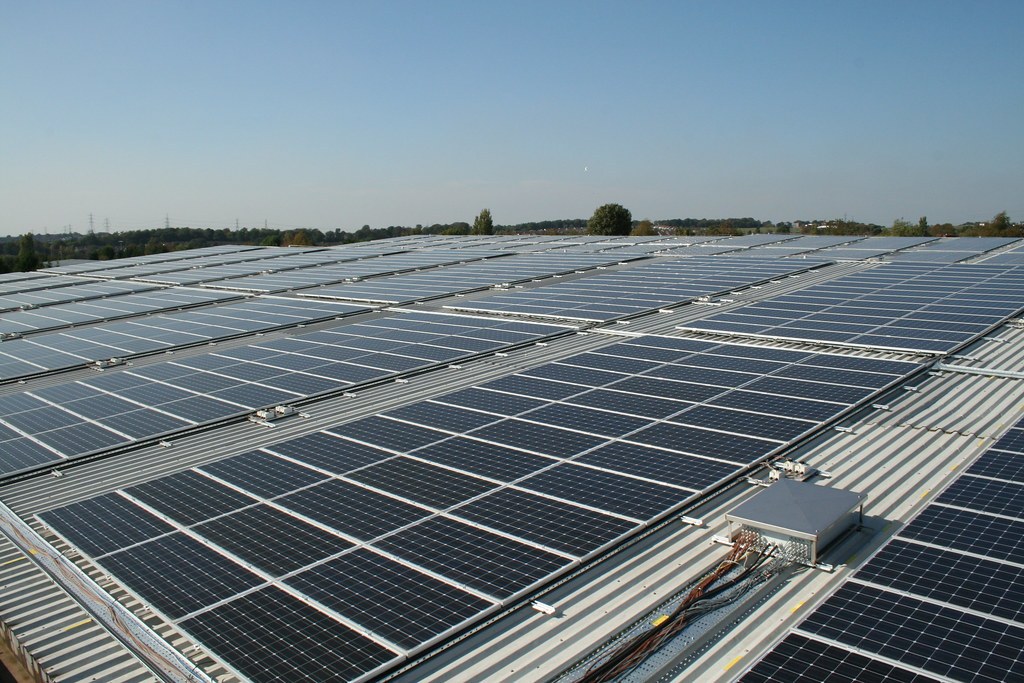 Aerial view of a vast rooftop solar panel array covering an industrial building, with trees and other rooftops visible in the distance